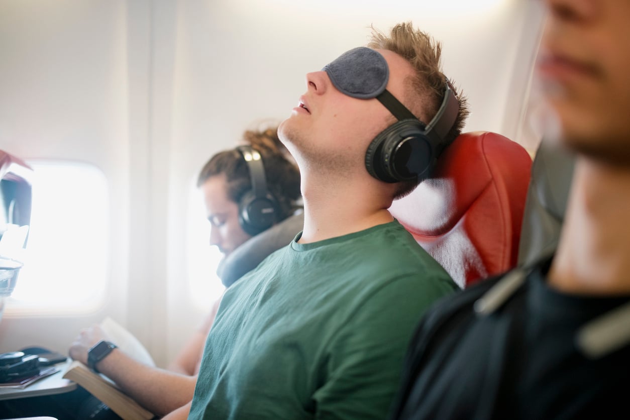 Man wearing headphones and a sleep mask rests on an airplane, seated between two other passengers; one is reading a book and the other is out of focus in the foreground.