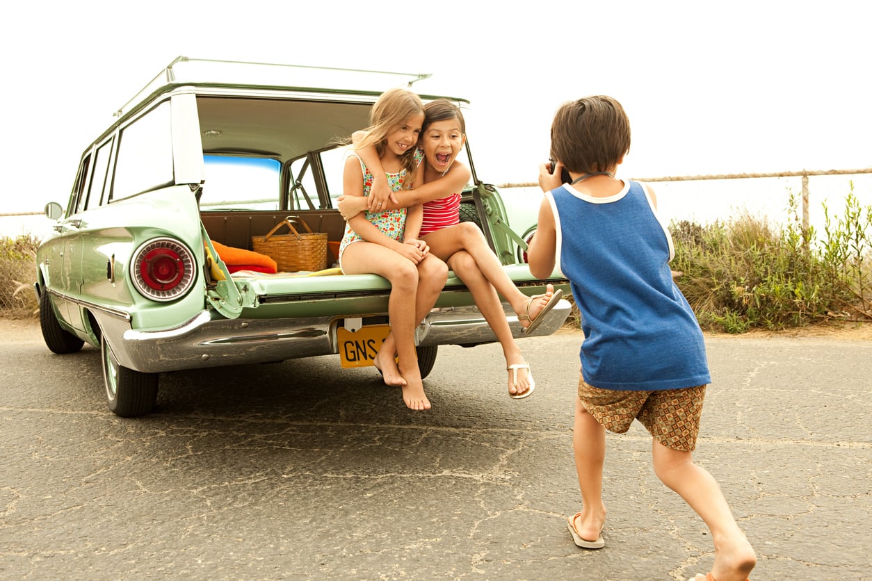 A boy takes a photo of two girls sitting on the open tailgate of a green vintage car. The children are smiling, and there is a picnic basket in the trunk. The scene appears cheerful and outdoorsy.