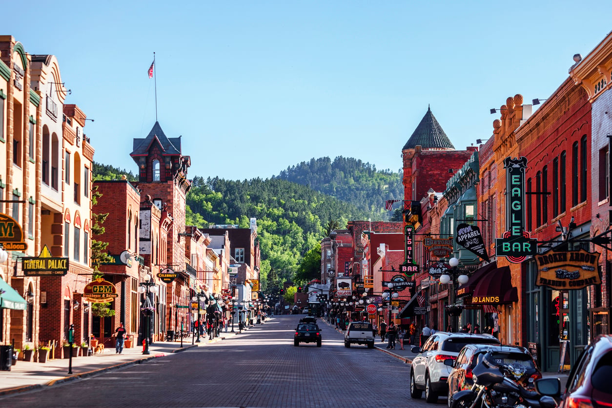 A lively historic downtown street with brick buildings, colorful signs, parked cars, and green hills in the background on a clear sunny day—one of the most charming nostalgic travel destinations for those seeking timeless small-town allure.