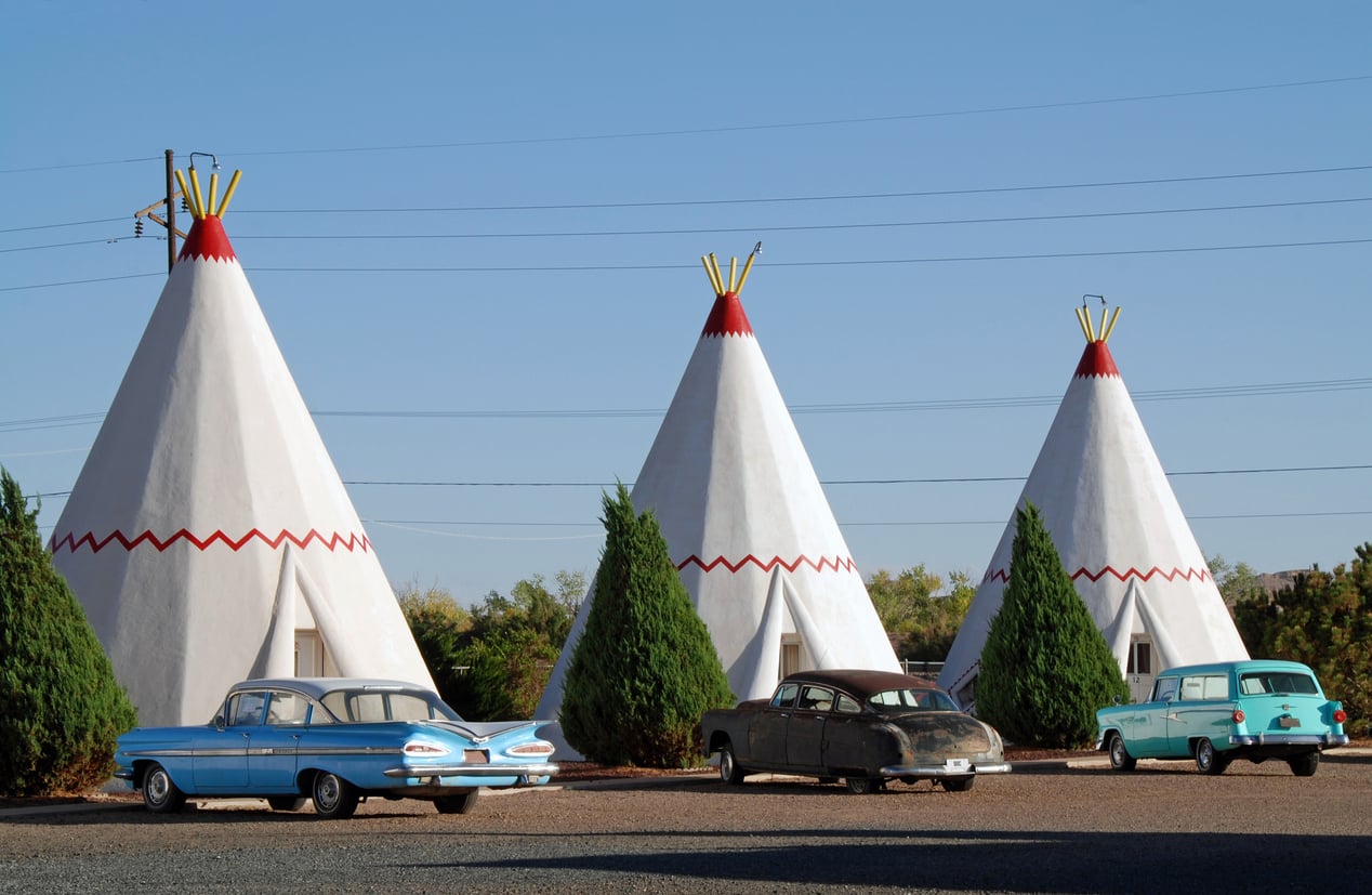 Three white teepee-shaped motel rooms with red zigzag trim, each parked with a classic car in front. Green shrubs and clear blue sky complete the retro roadside scene, capturing the timeless charm of Classic Roadside Attractions.