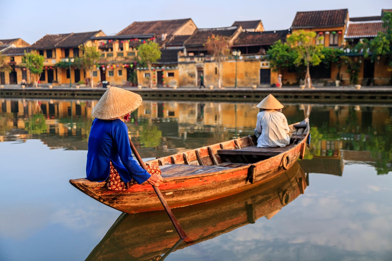 Two people wearing conical hats sit in a wooden boat on a calm river, with traditional yellow buildings and trees along the riverbank in the background—one of the travel spots boomers are learning to love for its timeless charm and tranquil scenery.