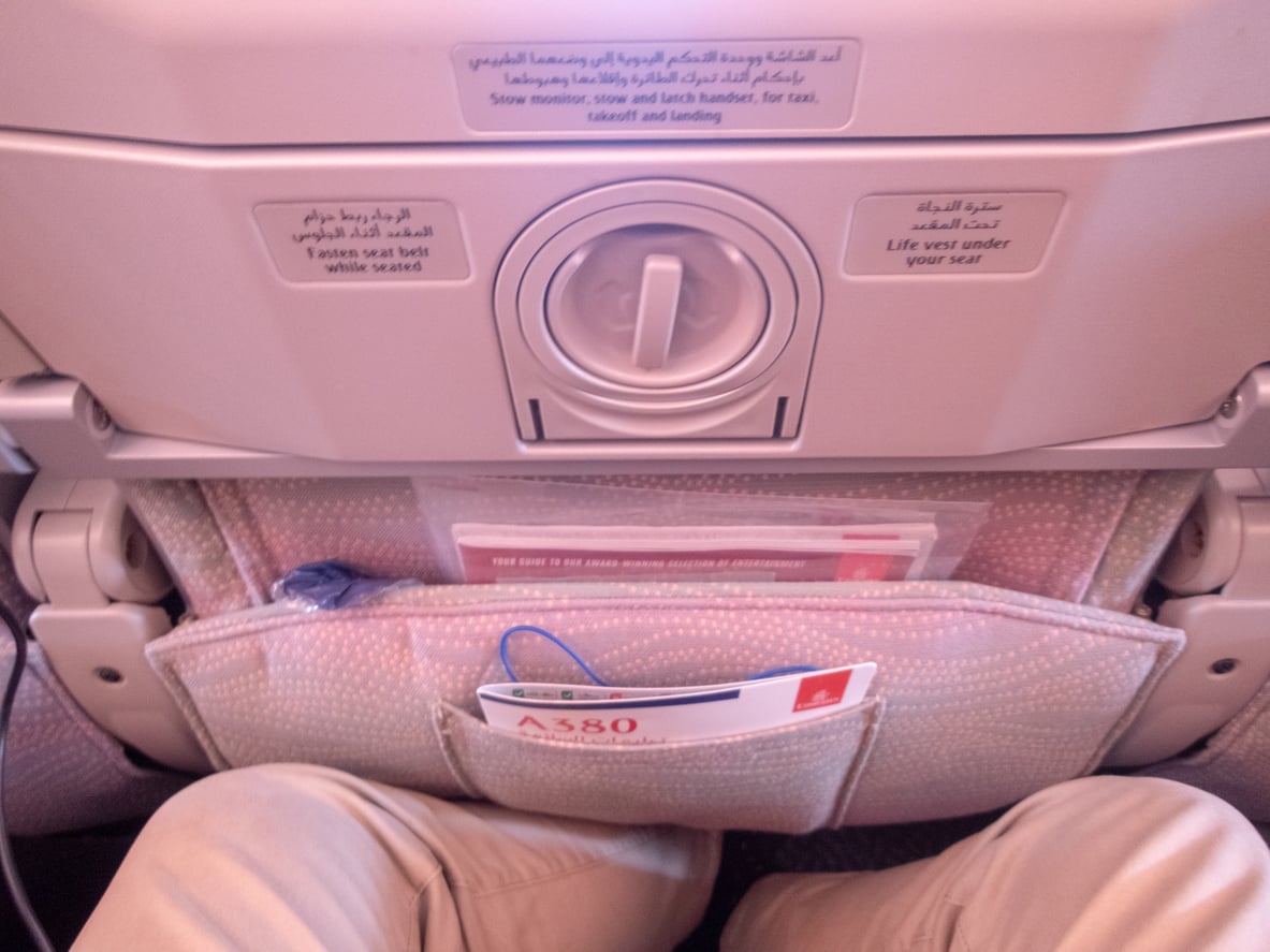 A person seated on an airplane with their legs visible, showing the back of the seat in front, a tray table, safety instructions, and a magazine in the seat pocket.