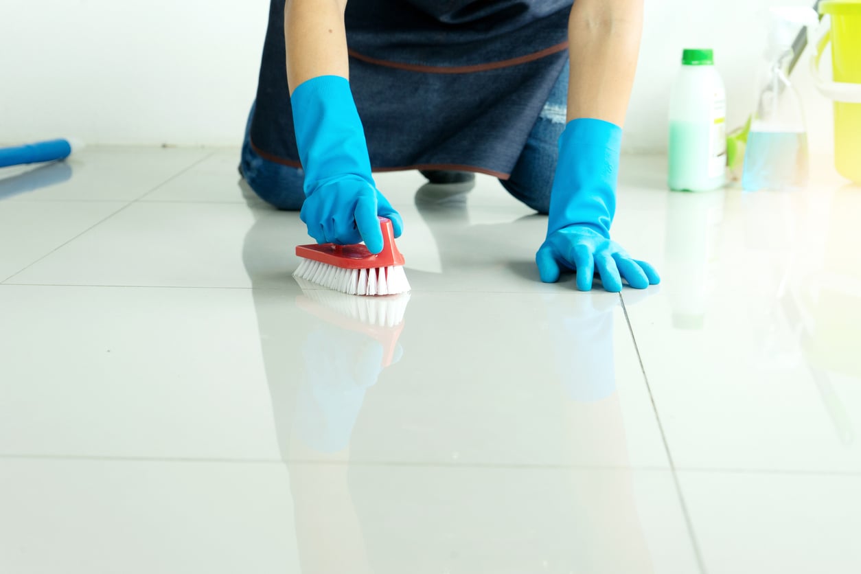 Person wearing blue gloves scrubs a shiny white tiled floor with a cleaning brush. Cleaning supplies, including bottles and a bucket, are visible in the background.