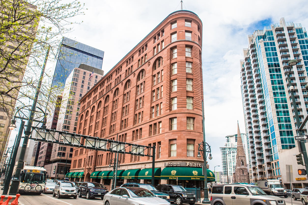 A historic red-brick hotel, recognized as one of the city’s esteemed Historic Hotels, stands proudly with its green awnings on a busy city street, surrounded by modern buildings, cars, and pedestrians under a partly cloudy sky.