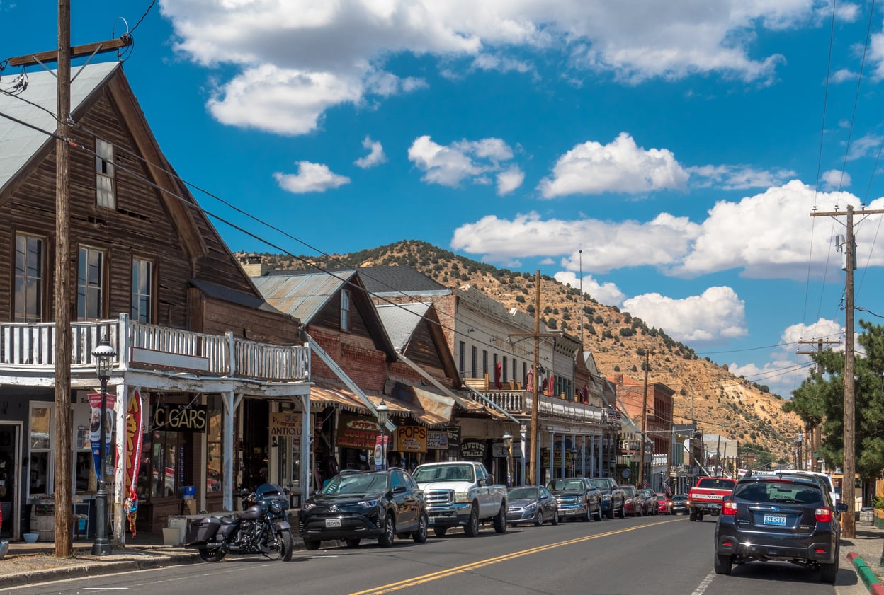 A western small town street with wooden storefronts, parked cars and motorcycles, and hills in the background under a blue sky with scattered clouds—one of those nostalgic travel destinations that invite you to step back in time.