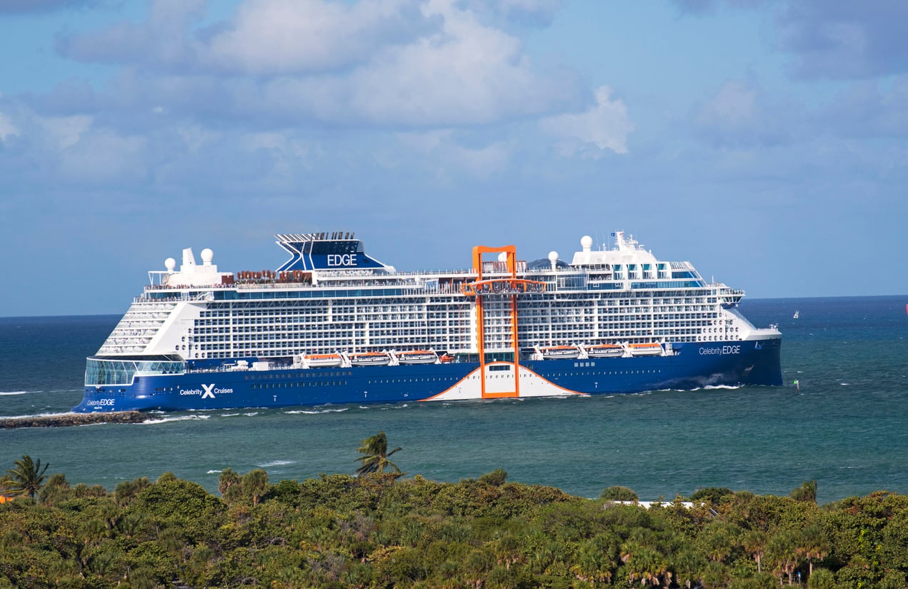 A large blue and white cruise ship from one of the renowned cruise lines sails near a green, tree-covered shoreline under a partly cloudy sky. The ship features several decks, large windows, and a distinctive orange structure on its side.