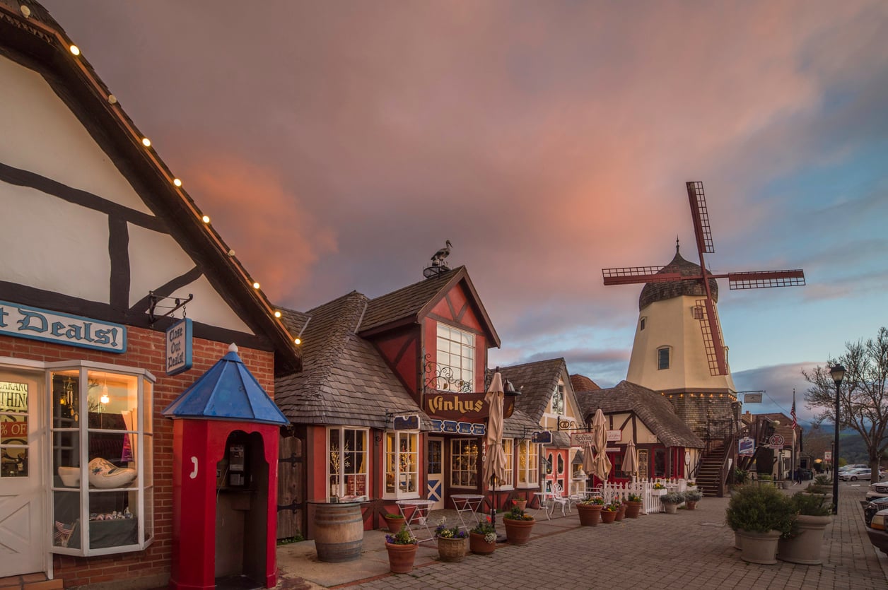 Colorful, old-fashioned storefronts line a quaint cobblestone street at sunset, with a tall windmill and dramatic clouds in the background, creating a charming village scene reminiscent of nostalgic travel destinations.