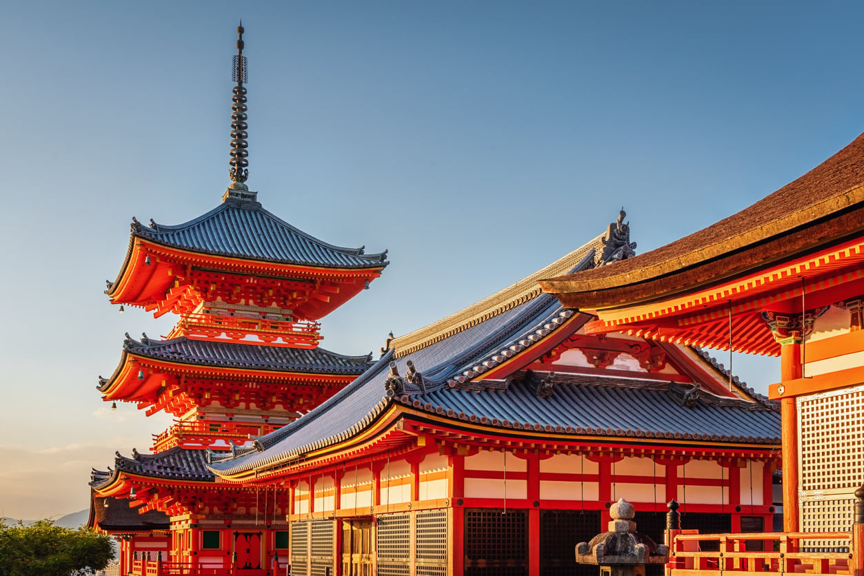 Traditional Japanese temple buildings with vibrant red and white colors, curved tiled roofs, and a tall pagoda under a clear blue sky in warm sunlight.