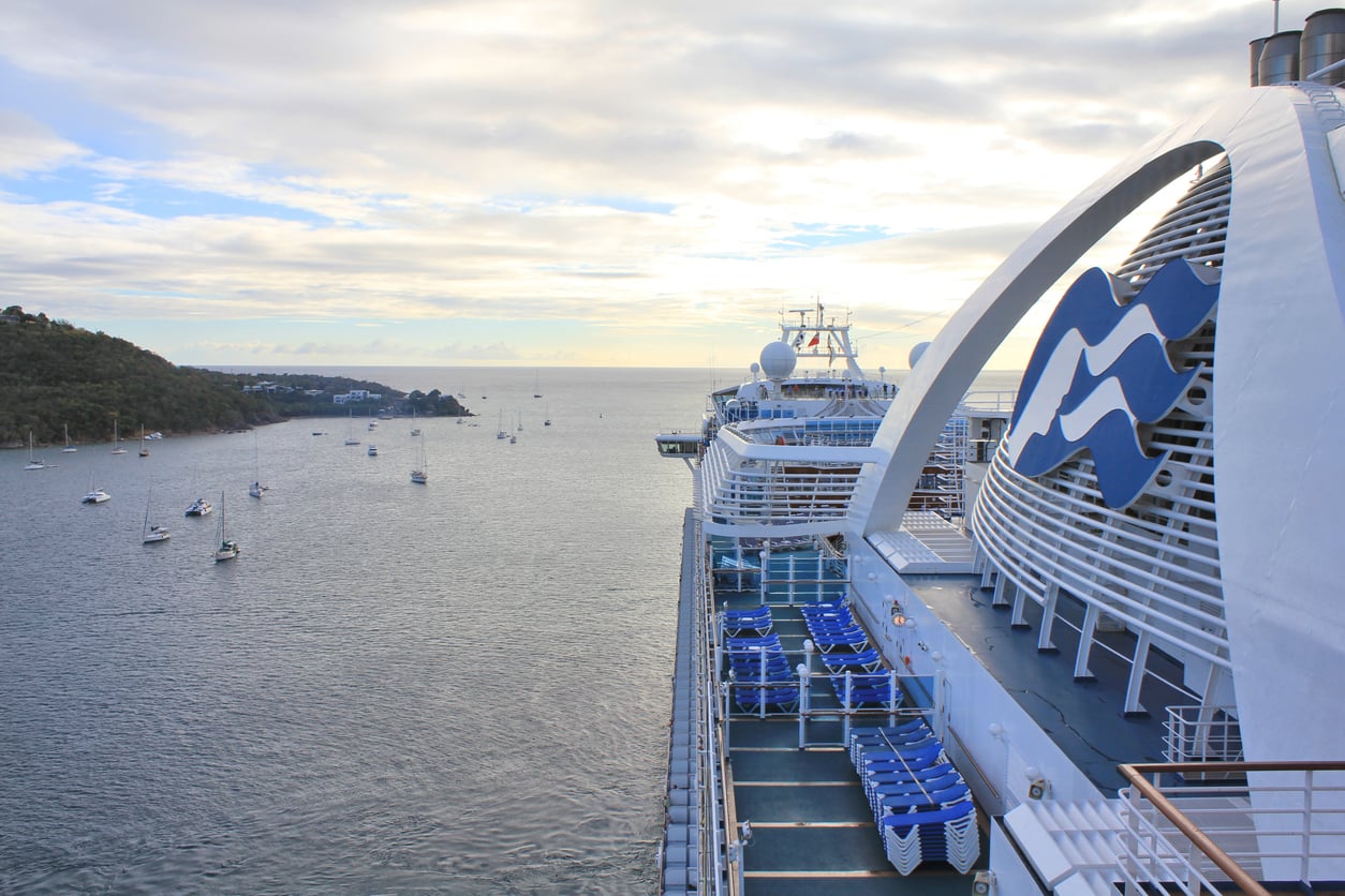 A large cruise ship with blue lounge chairs is docked near a calm bay. Several small boats float in the water under a partly cloudy sky, with a green hillside on the left.
