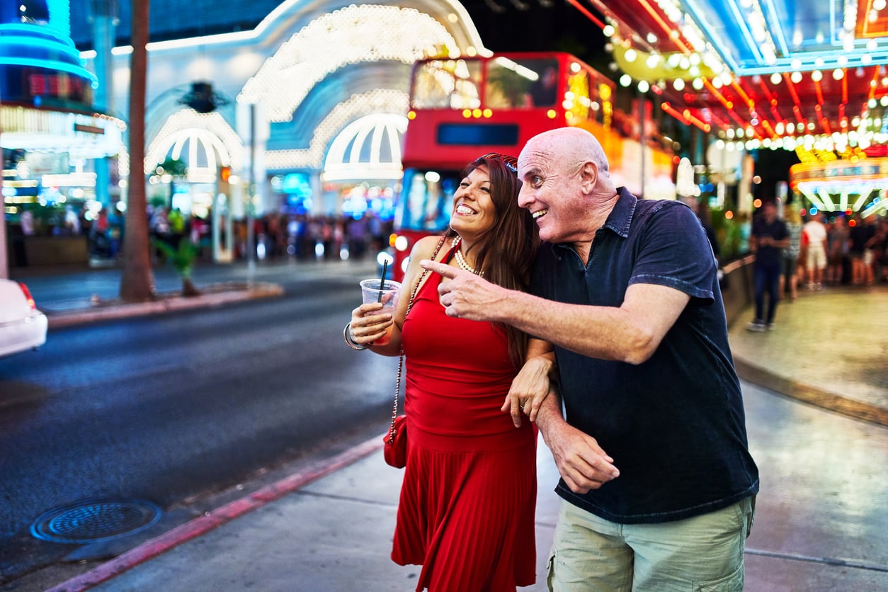 A smiling couple walk arm in arm under bright neon lights on a lively city street at night, exploring travel destinations boomers are skipping. The woman in a red dress holds a drink while the man in a dark shirt points excitedly at something ahead.