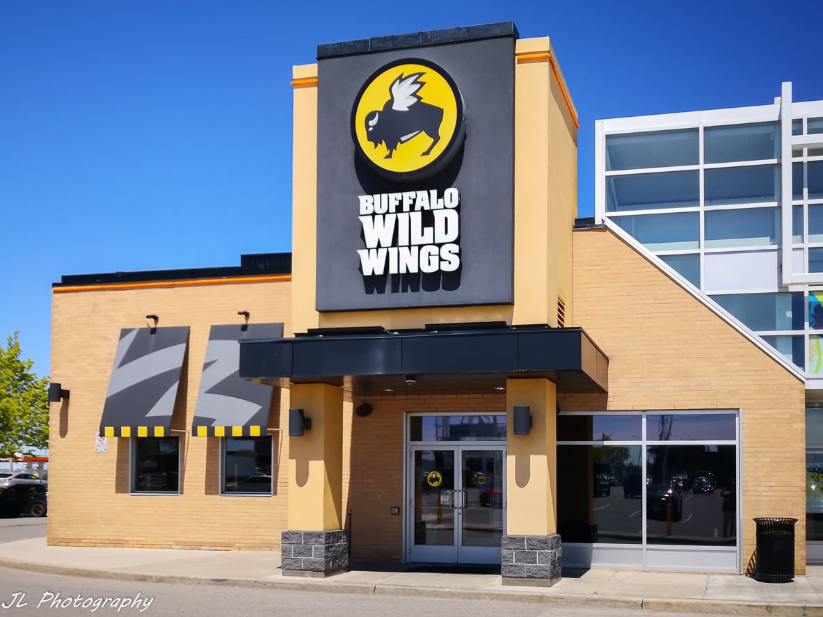 A Buffalo Wild Wings restaurant with a yellow and black sign featuring a buffalo logo on the tan building exterior, sunny day, glass windows, and a parking lot in front.