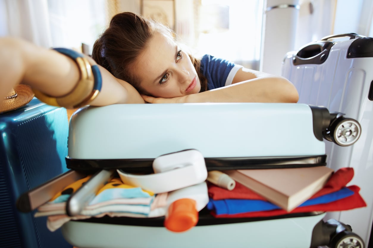 A woman rests her head on an overstuffed suitcase, looking tired and frustrated. Clothes, a book, and toiletries spill out of the suitcase, with more luggage seen in the background.