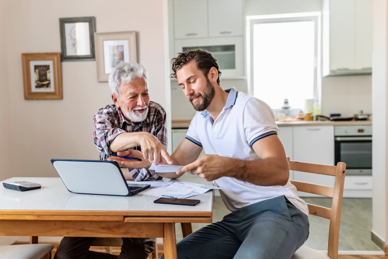 Two men sit at a kitchen table with a laptop and papers, smiling and discussing documents. One points at the paper while the other holds it. The setting is bright and homey.