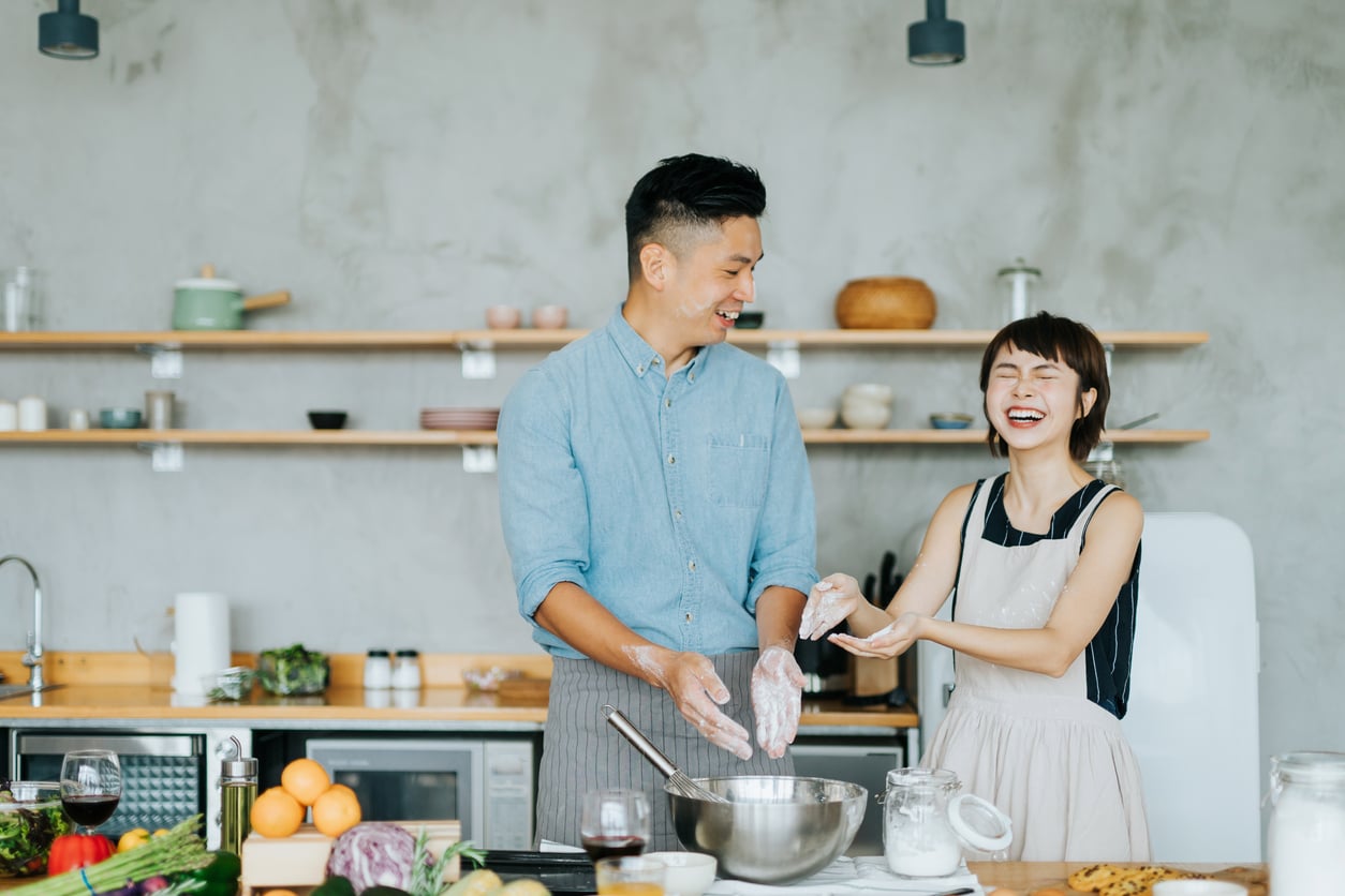 A man and woman laugh together in a modern kitchen while preparing dough. Their hands are covered in flour, and fresh vegetables and baking ingredients are on the counter.
