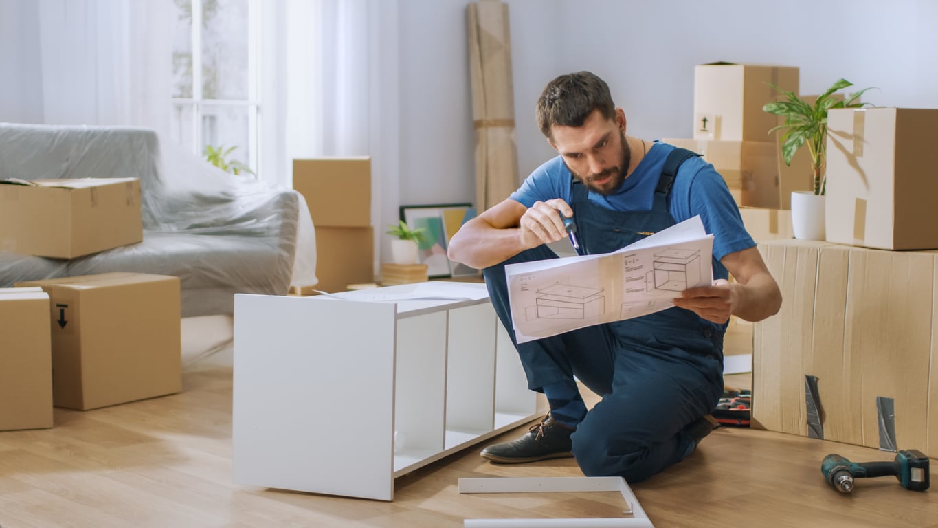 A man in overalls kneels on the floor, reading assembly instructions while assembling white furniture. Cardboard boxes and tools are scattered around a room with a couch and potted plants.