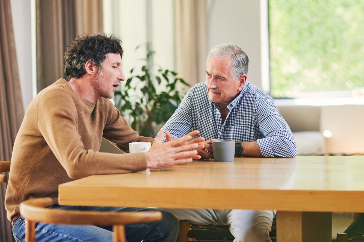 Two men sit at a wooden table with coffee mugs, engaged in a serious conversation. One gestures with his hands while the other listens attentively. Natural light fills the room through large windows.