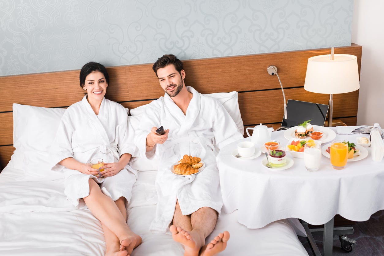 A smiling couple in white bathrobes sits on a hotel bed with breakfast foods and drinks on a table. Reminiscent of things hotels used to offer guests, the cozy setting exudes comfort, as one person holds a TV remote and both appear relaxed, enjoying their meal.