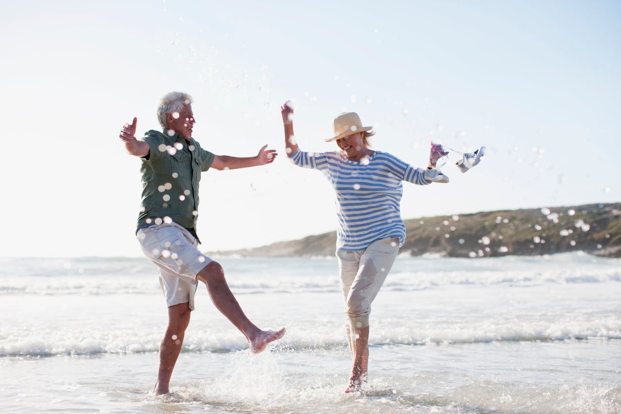 Two older adults joyfully splash water at each other on a sunny beach, smiling and laughing. One person wears a hat and striped shirt; the other wears a short-sleeve shirt and shorts. Waves roll in behind them.