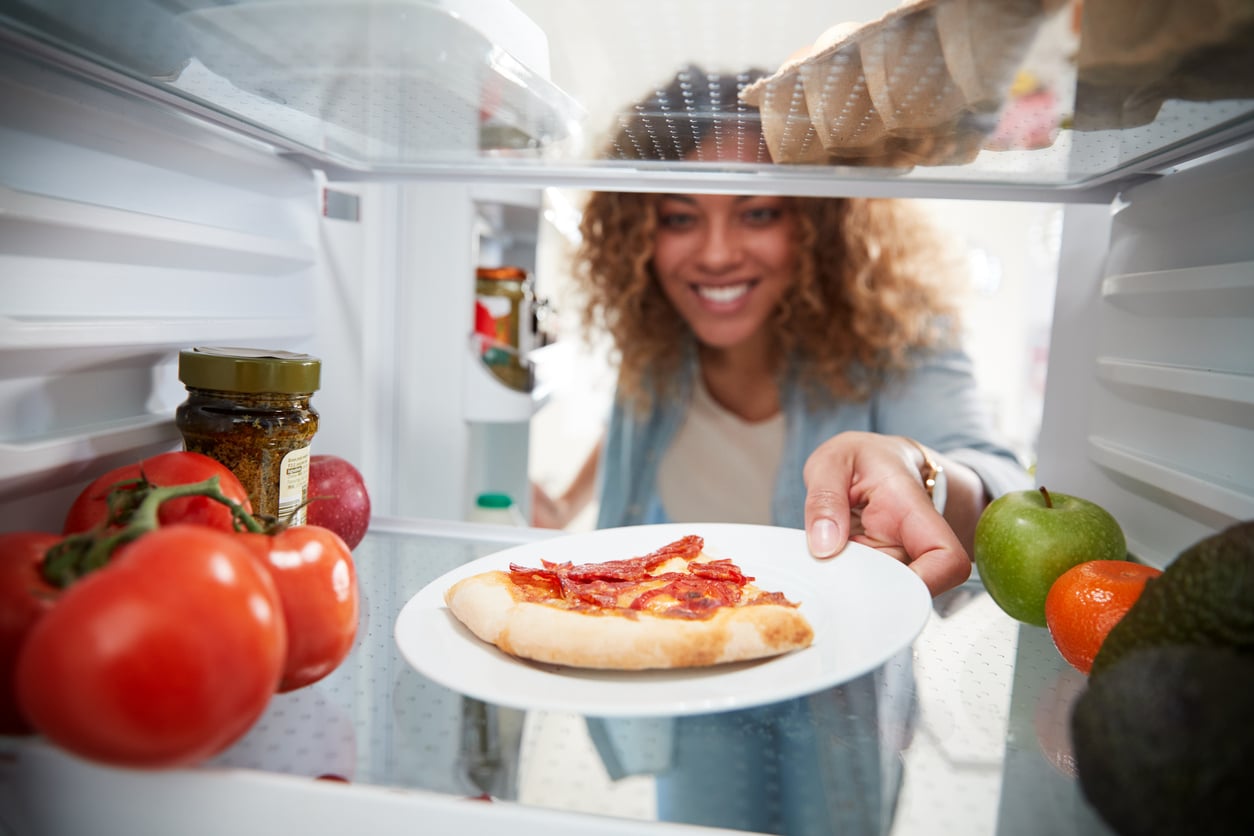 A woman smiling as she places a plate with a slice of pepperoni pizza into a refrigerator, surrounded by fresh fruits and vegetables.