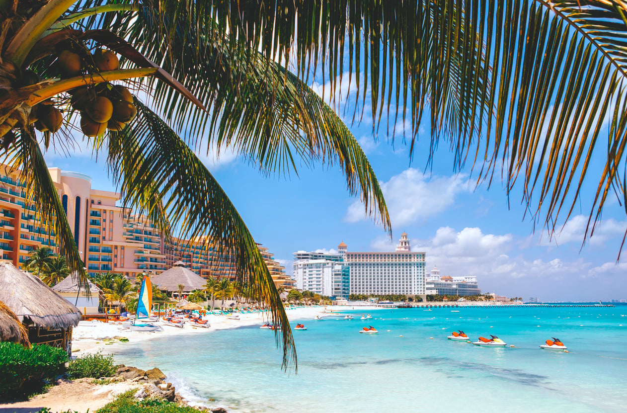 A tropical beach with turquoise water, palm trees, hotels, jet skis, and people enjoying the sunny day under a bright blue sky—despite some complaints about the bad food served at nearby beachside cafes.