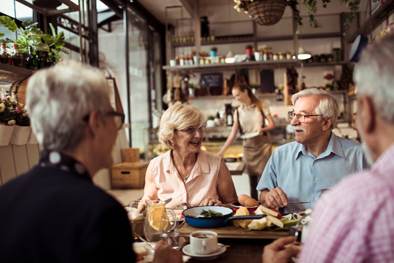 Four older adults sit around a table in a bright, cozy cafe, enjoying food and drinks together while chatting and smiling. A server is seen in the background carrying a tray of food.