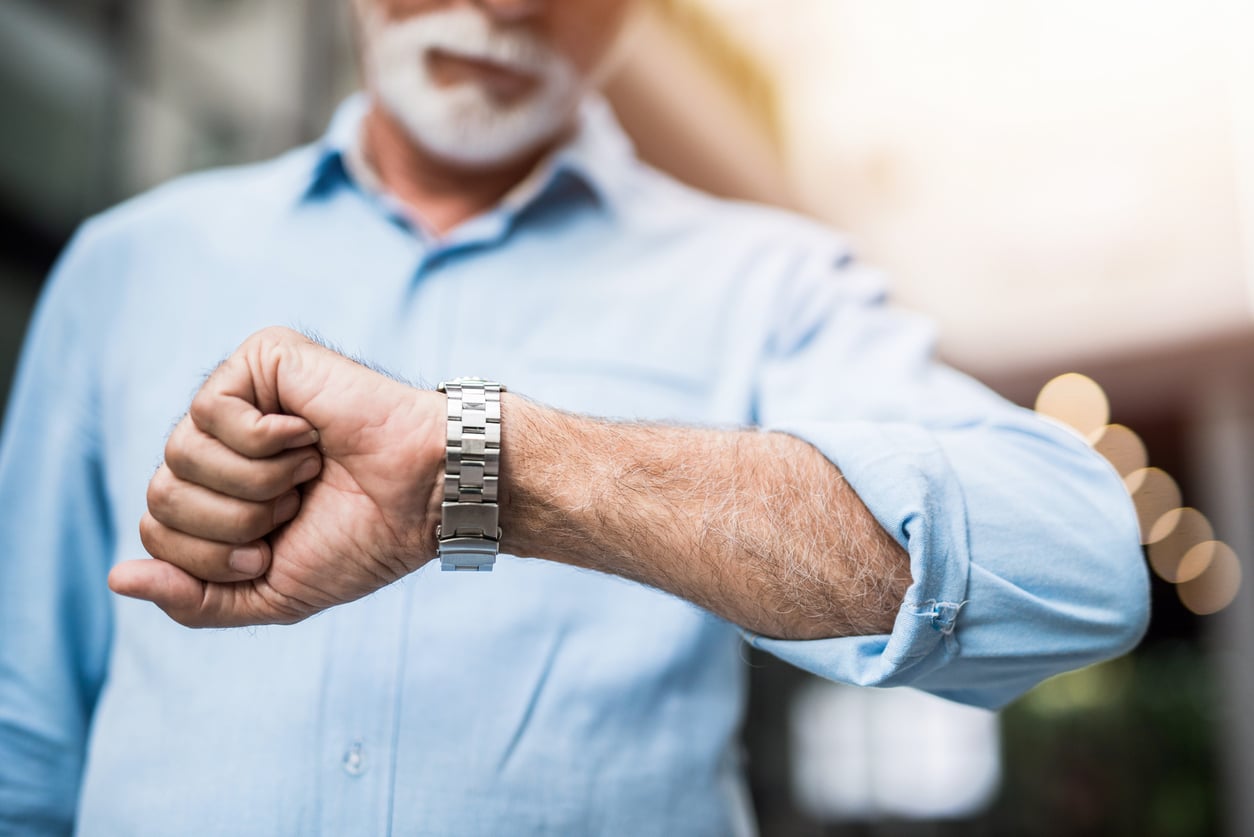 A man with a white beard in a blue shirt looks at his wristwatch, appearing to check the time. The background is blurred, focusing attention on his watch and arm.