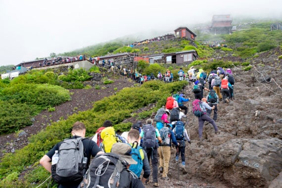 Hikers with backpacks ascend a rocky, steep trail on a foggy mountain, with green shrubbery and several buildings visible ahead.