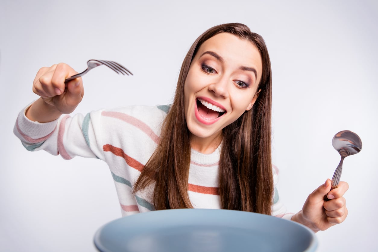 Smiling woman holding a fork and spoon looks eagerly at an empty blue bowl in front of her, appearing excited and ready to eat.