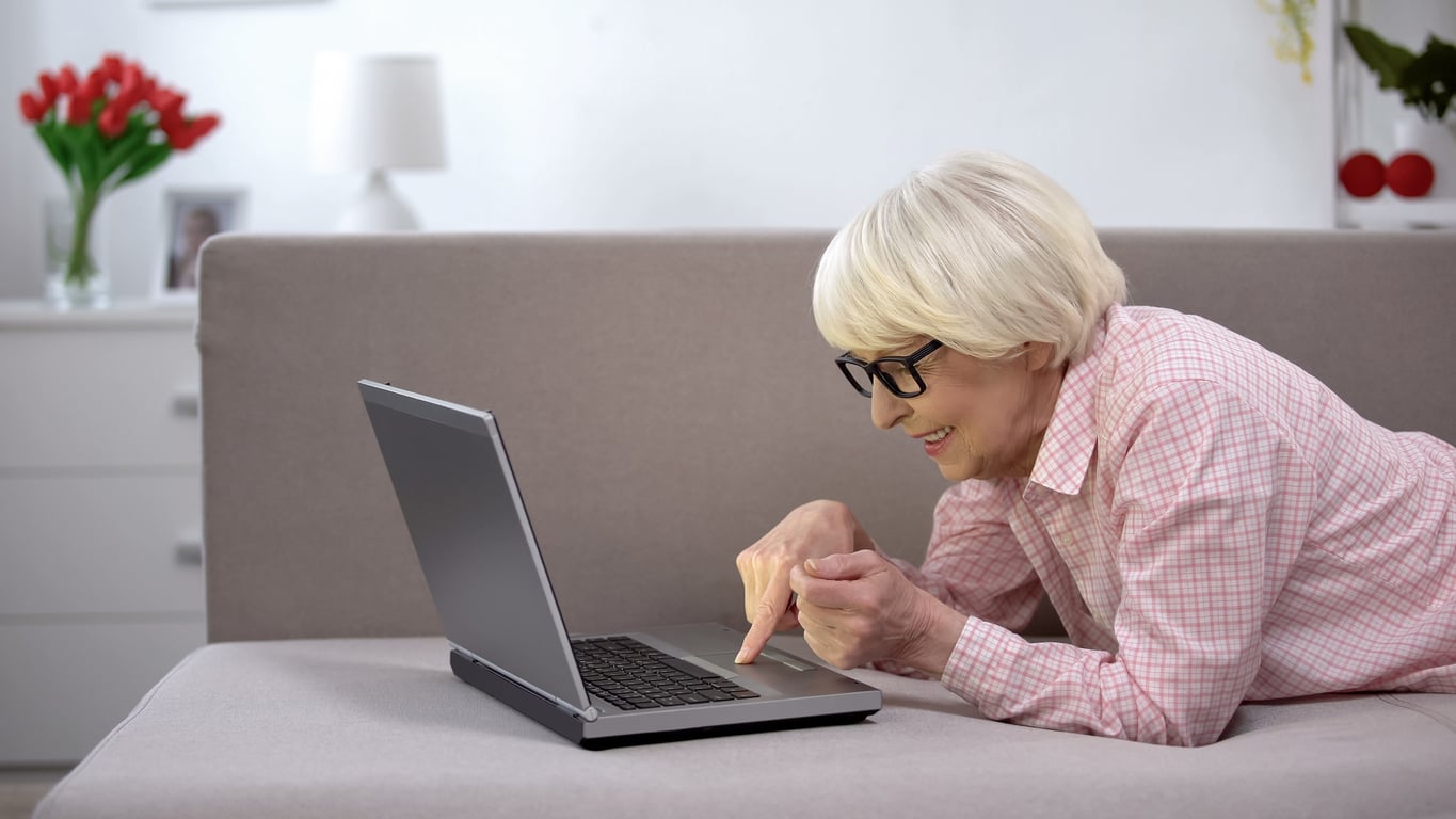 An older woman with short white hair and glasses is lying on a couch, smiling while using a laptop. She is wearing a pink plaid shirt. A white dresser and red flowers are visible in the background.