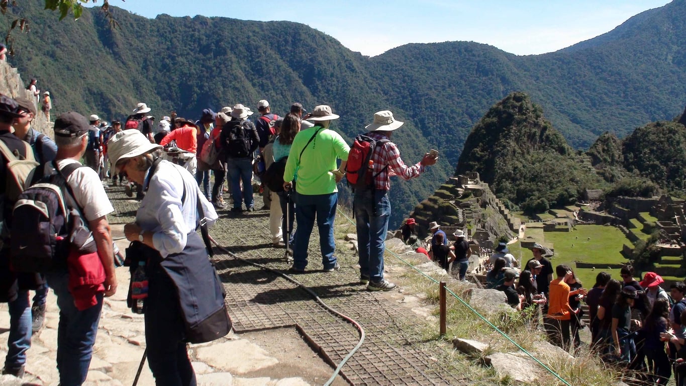 Tourists wearing hats and backpacks stand on a pathway overlooking ancient stone ruins at Machu Picchu, with green mountains and blue sky in the background.