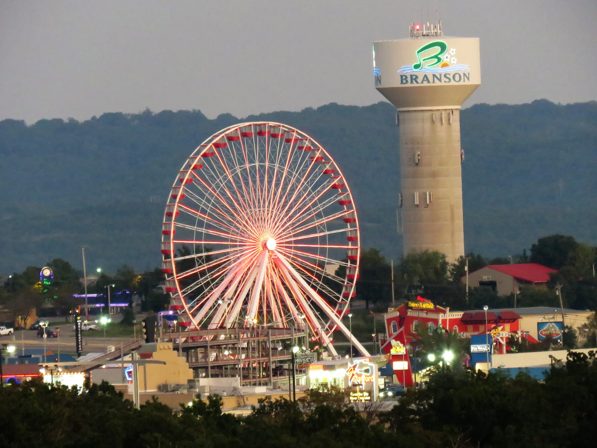 A large Ferris wheel lit up at dusk stands near a tall water tower labeled “Branson,” creating a picturesque scene perfect for exploring after age 50, with rolling hills and various buildings in the background.