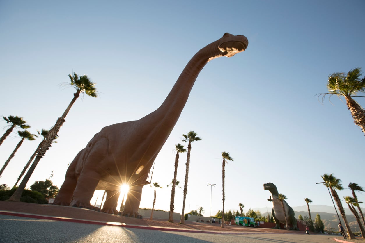 Two large dinosaur statues, reminiscent of classic roadside attractions, stand among palm trees under a clear blue sky. The sun is near the horizon, shining through the legs of the nearest dinosaur and casting long shadows on the ground.