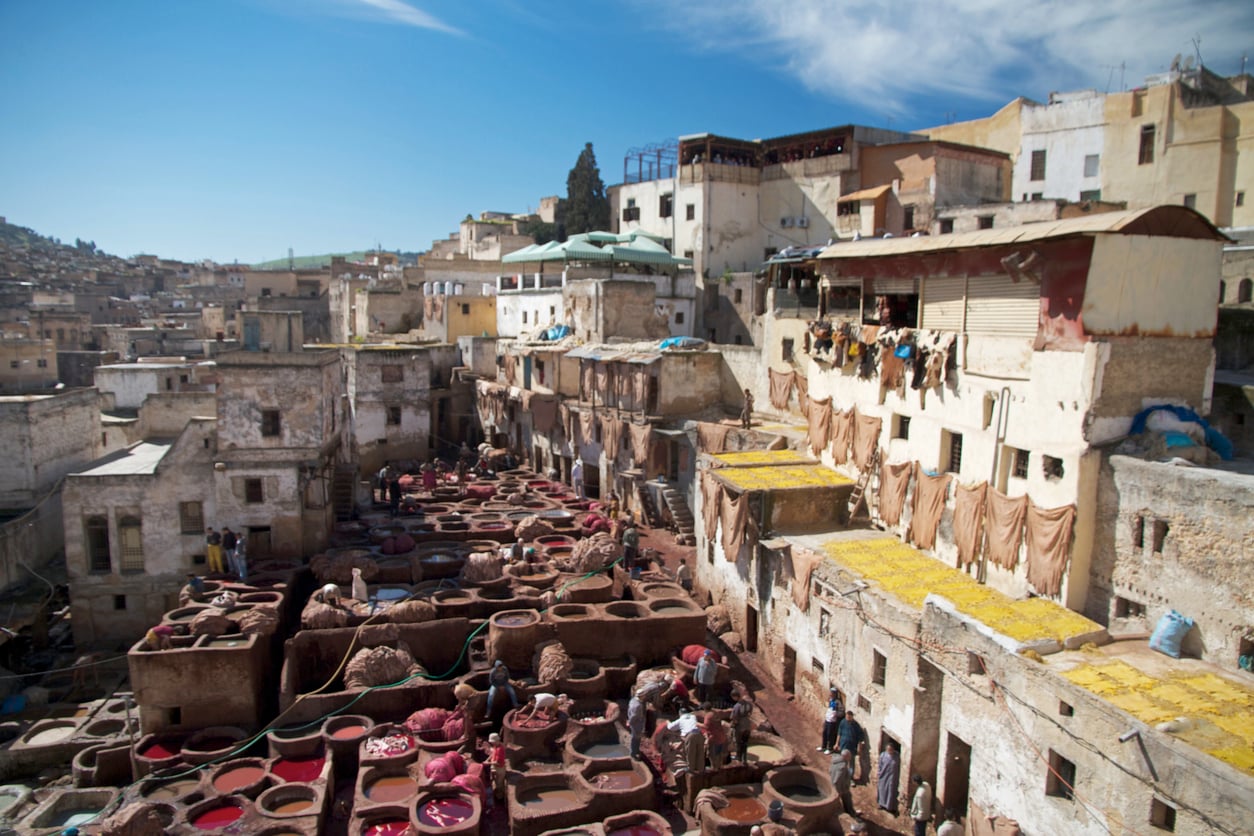View of a traditional tannery in a historic Moroccan city, with workers among large vats of colorful dyes and old buildings surrounding the area under a blue sky.