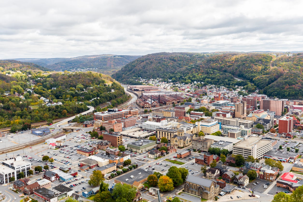 Aerial view of a small city with clustered buildings, roads, and parking lots, surrounded by green, tree-covered hills under a cloudy sky.