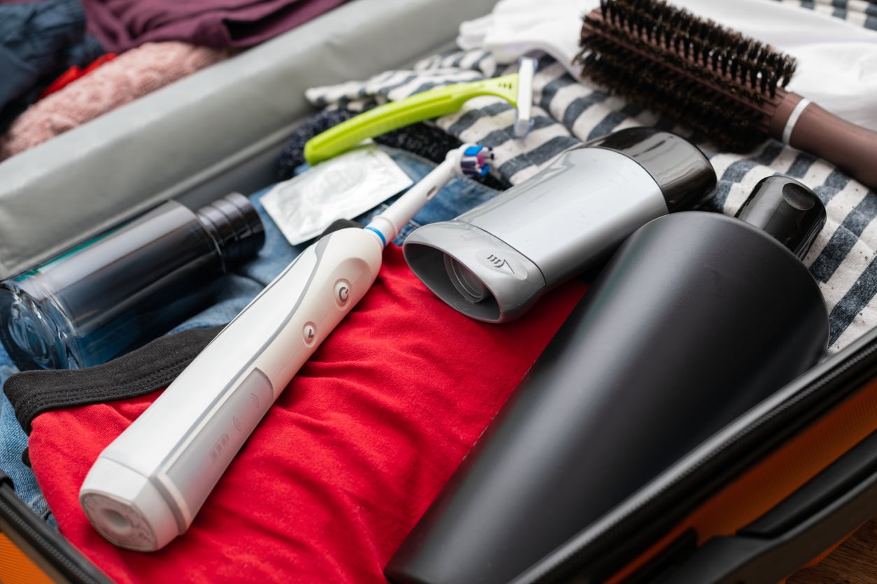 An open suitcase with clothes, an electric toothbrush, a hair dryer, a water bottle, a round hairbrush, a razor, and a condom on top.