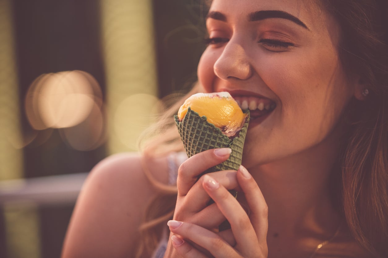 A woman smiling and about to eat an ice cream cone with orange and white scoops, holding it with both hands. The background is softly blurred with warm lighting.