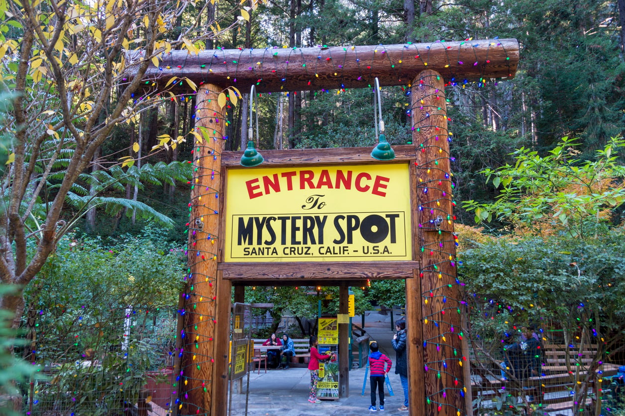 A wooden entrance sign reads “Entrance to Mystery Spot, Santa Cruz, Calif., U.S.A.” with people walking underneath, surrounded by trees and string lights—capturing the nostalgic charm of classic roadside attractions.