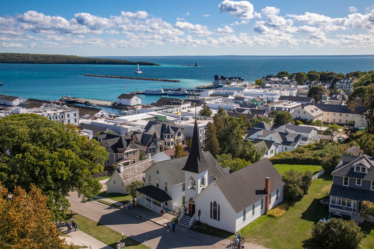 Aerial view of a coastal town with a church in the foreground, houses, trees, a marina, and blue water under a partly cloudy sky—one of those nostalgic travel destinations that evoke memories of tranquil seaside escapes.
