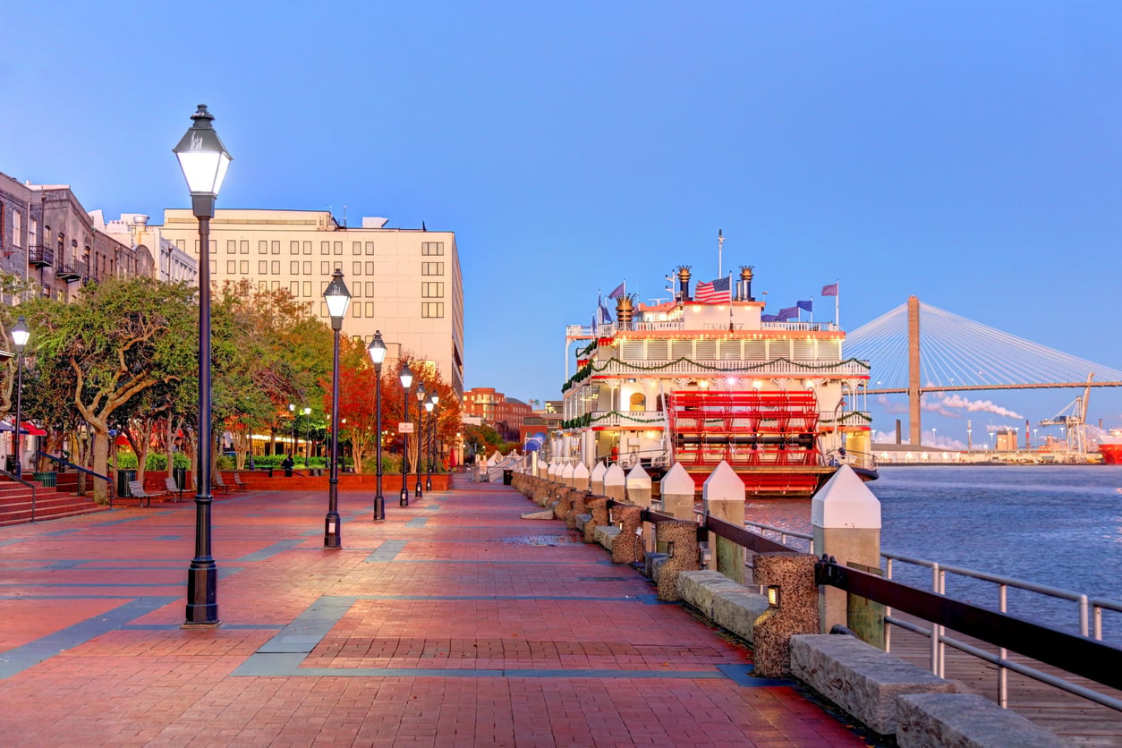 A brick walkway along a river with street lamps, trees, and a large riverboat docked evokes the charm of nostalgic travel destinations. Buildings and a cable-stayed bridge are visible in the background under a clear sky.