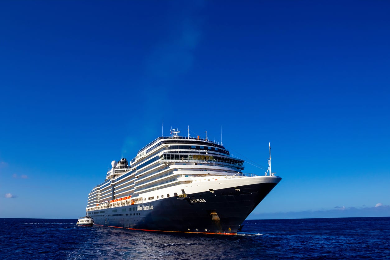 A large white and navy cruise ship from one of the world’s leading cruise lines sails on calm blue ocean water under a clear blue sky, with another smaller boat visible near its side.