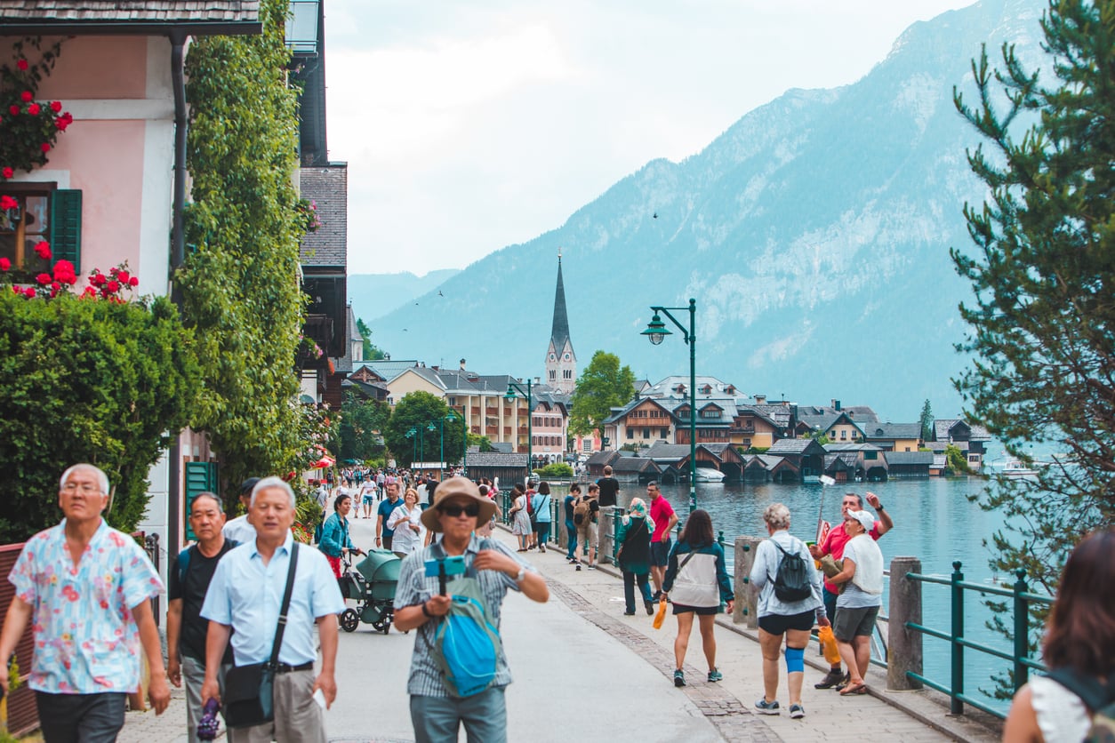Tourists walk along a lakeside promenade lined with houses and greenery, with mountains and a church steeple visible in the background under a partly cloudy sky.