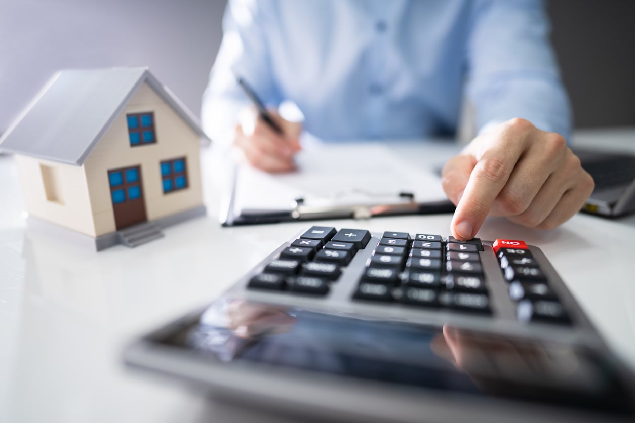 A person presses buttons on a calculator next to a small model house, writing on paper, suggesting calculations related to real estate or mortgage planning.