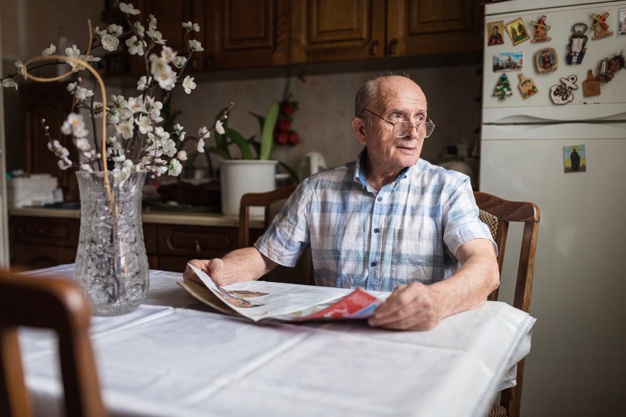 An elderly man wearing glasses and a plaid shirt sits at a kitchen table with a newspaper. A glass vase with flowers is on the table, and a refrigerator with magnets is visible in the background.