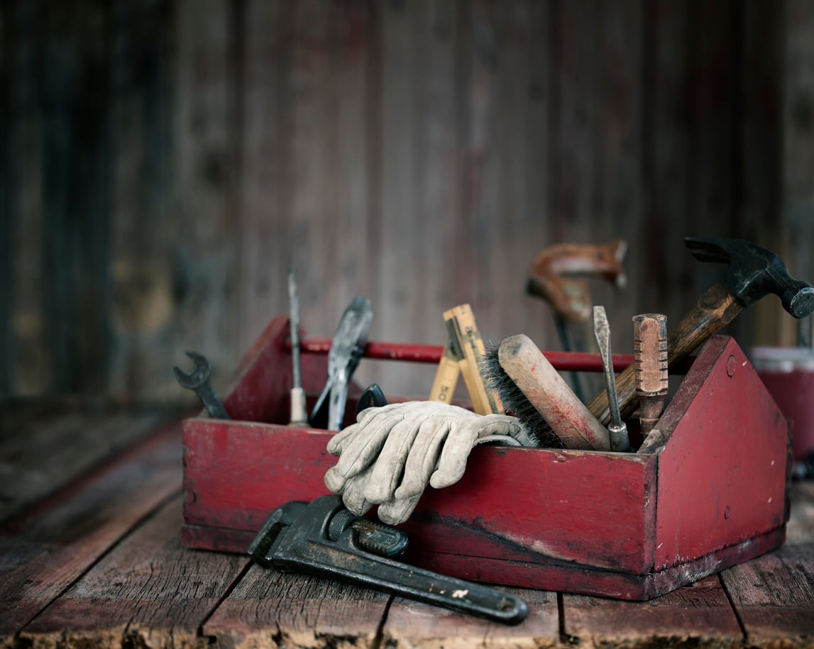 A weathered red toolbox sits on a wooden surface, filled with assorted hand tools and a pair of work gloves draped across the top. The scene has a rustic, vintage feel with a blurred wooden background.