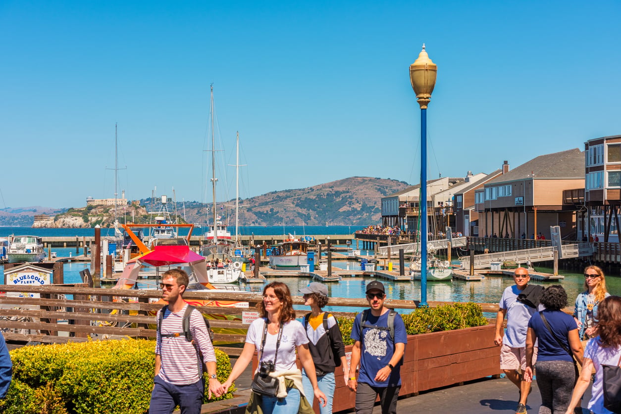 People walk along a sunny waterfront with boats docked at a marina, buildings, and a distant island with hills in the background under a clear blue sky.