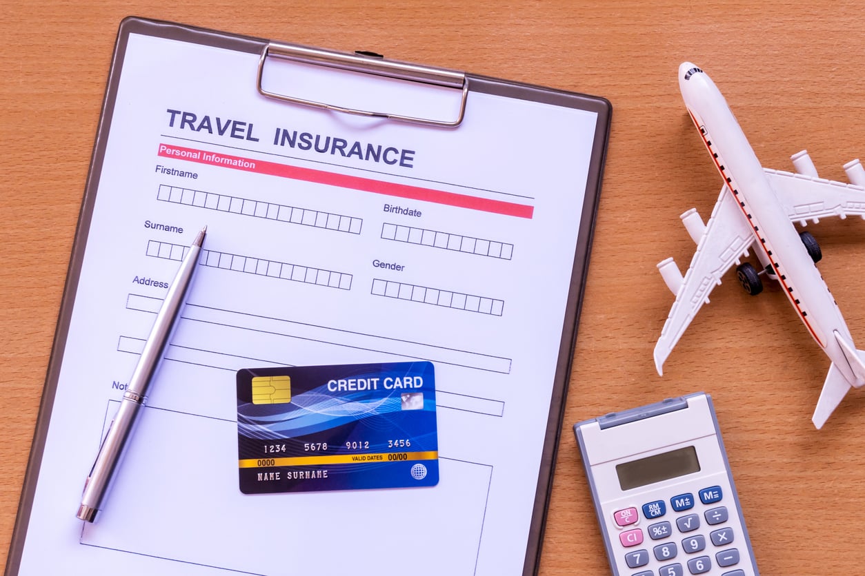 A travel insurance form on a clipboard, with a pen, credit card, toy airplane, and calculator on a wooden surface.