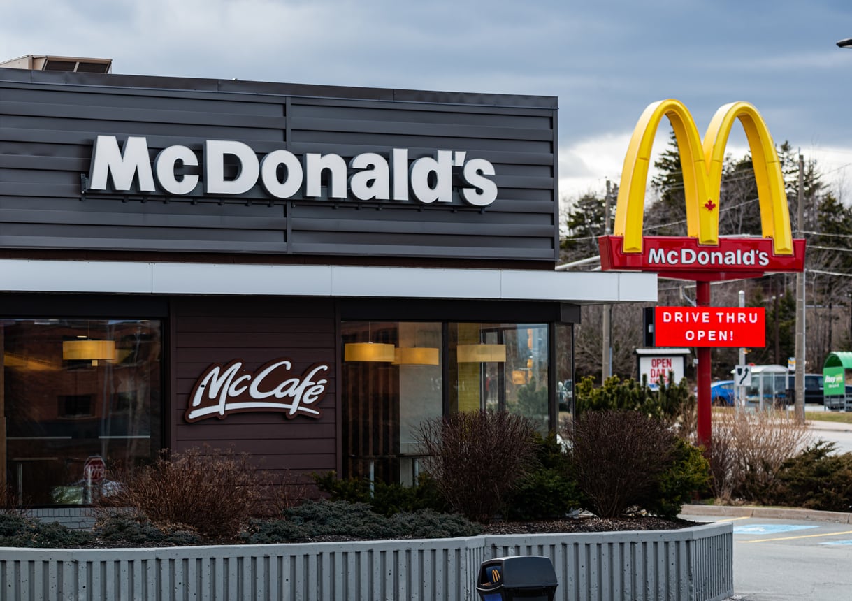 A McDonald’s restaurant with a McCafé sign, large golden arches, and a lit sign that reads “Drive Thru Open!” Bushes and trees surround the building under a cloudy sky.