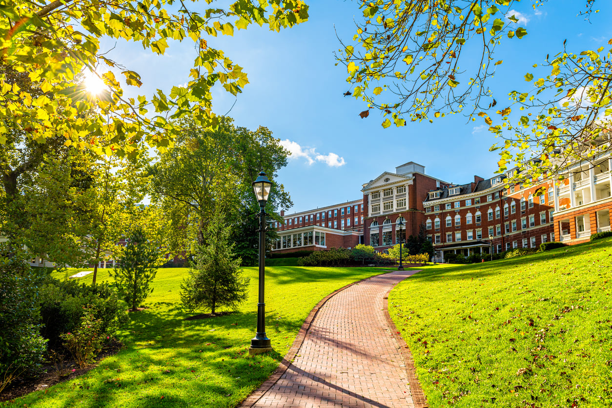 A red brick path leads through a green, sunny campus lawn toward a large red brick academic building with white trim, reminiscent of the timeless architecture found at many Historic Hotels, and is surrounded by leafy trees under a clear blue sky.