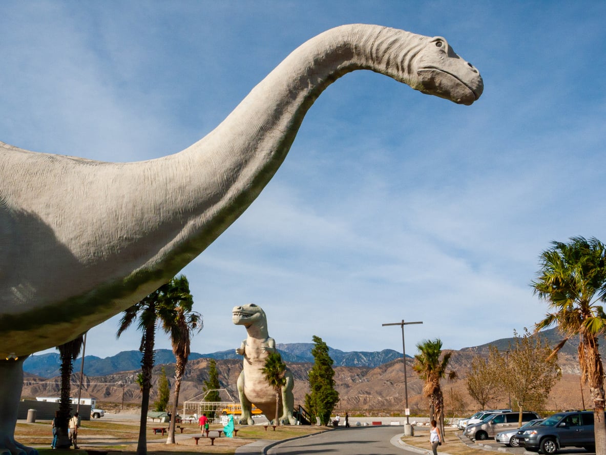 Two giant dinosaur statues stand among palm trees near a road with people walking around, set against a backdrop of mountains and a blue sky.