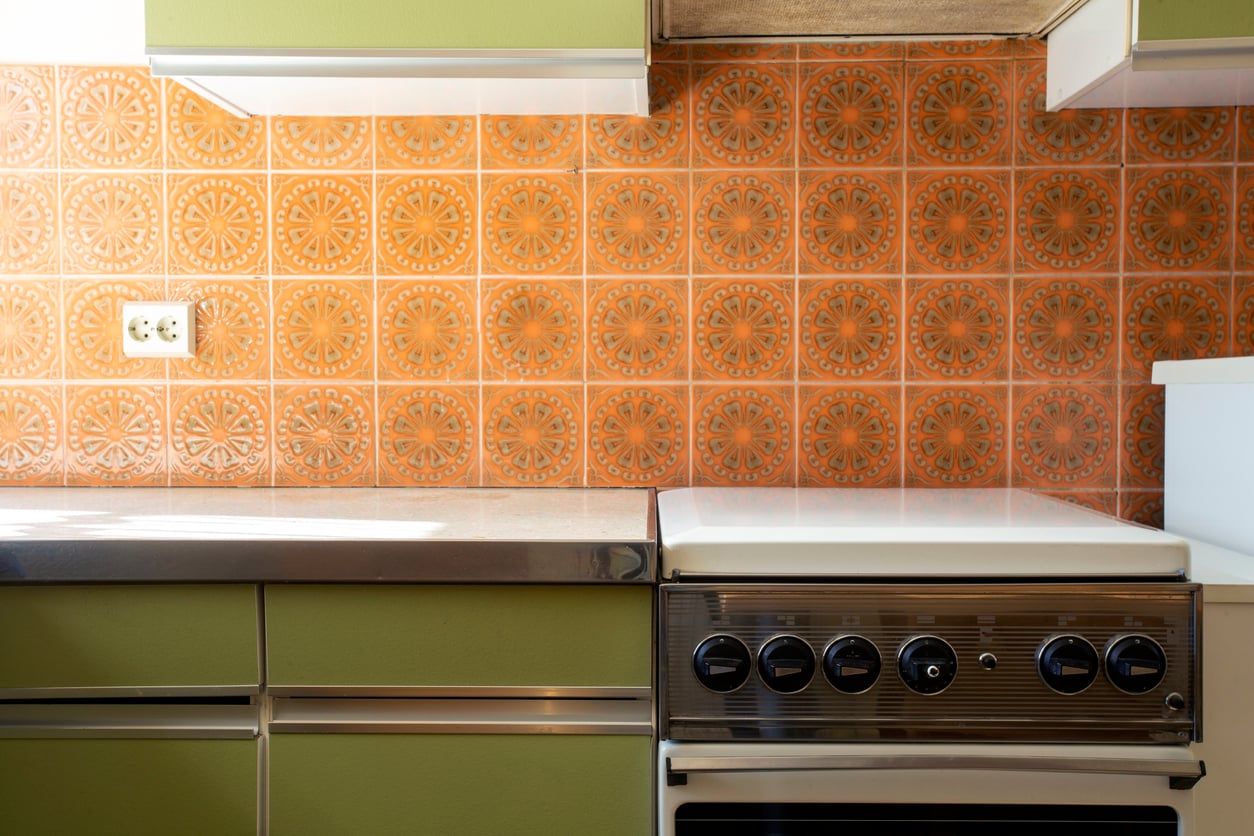 Vintage kitchen with olive green cabinets, orange patterned tile backsplash, stainless steel countertop, and an old-fashioned stove with black knobs.